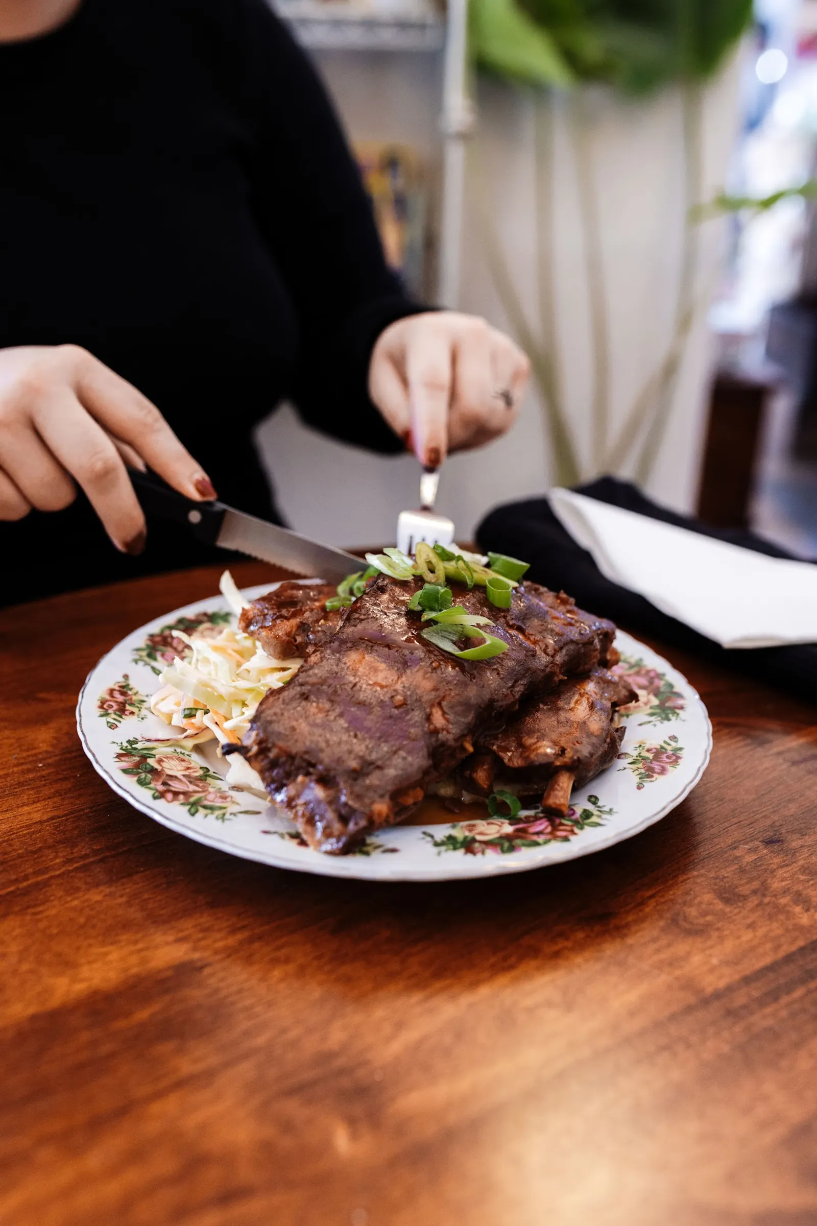 Slow-cooked pork ribs on vintage floral plate with coleslaw and spring onions, being cut by a diner